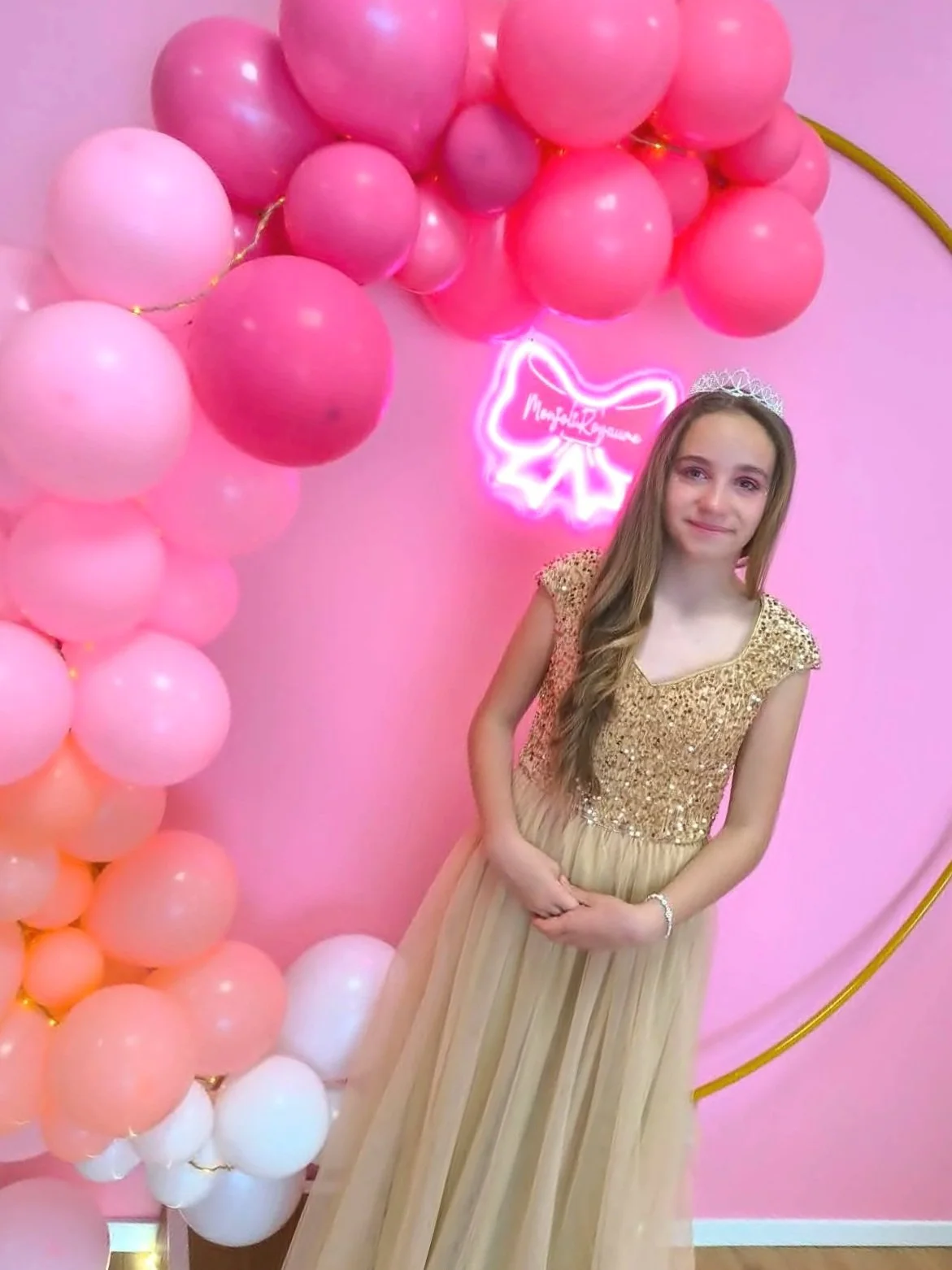 Jeune fille habillée en robe dorée avec une couronne de princesse, posant devant une décoration de ballons roses et lilas, avec un éclairage neon rose en forme de papillon en arrière-plan.