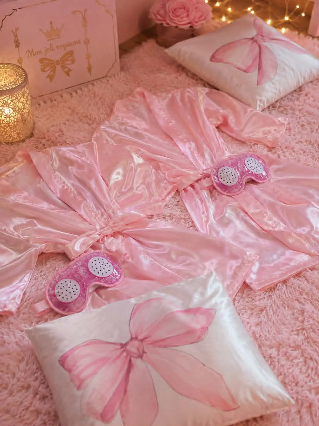 Décor de chambre rose avec coussins à motifs de papillon, robe en satin rose, masques de sommeil en plastique rose, et ambiance chaleureuse avec guirlandes lumineuses.