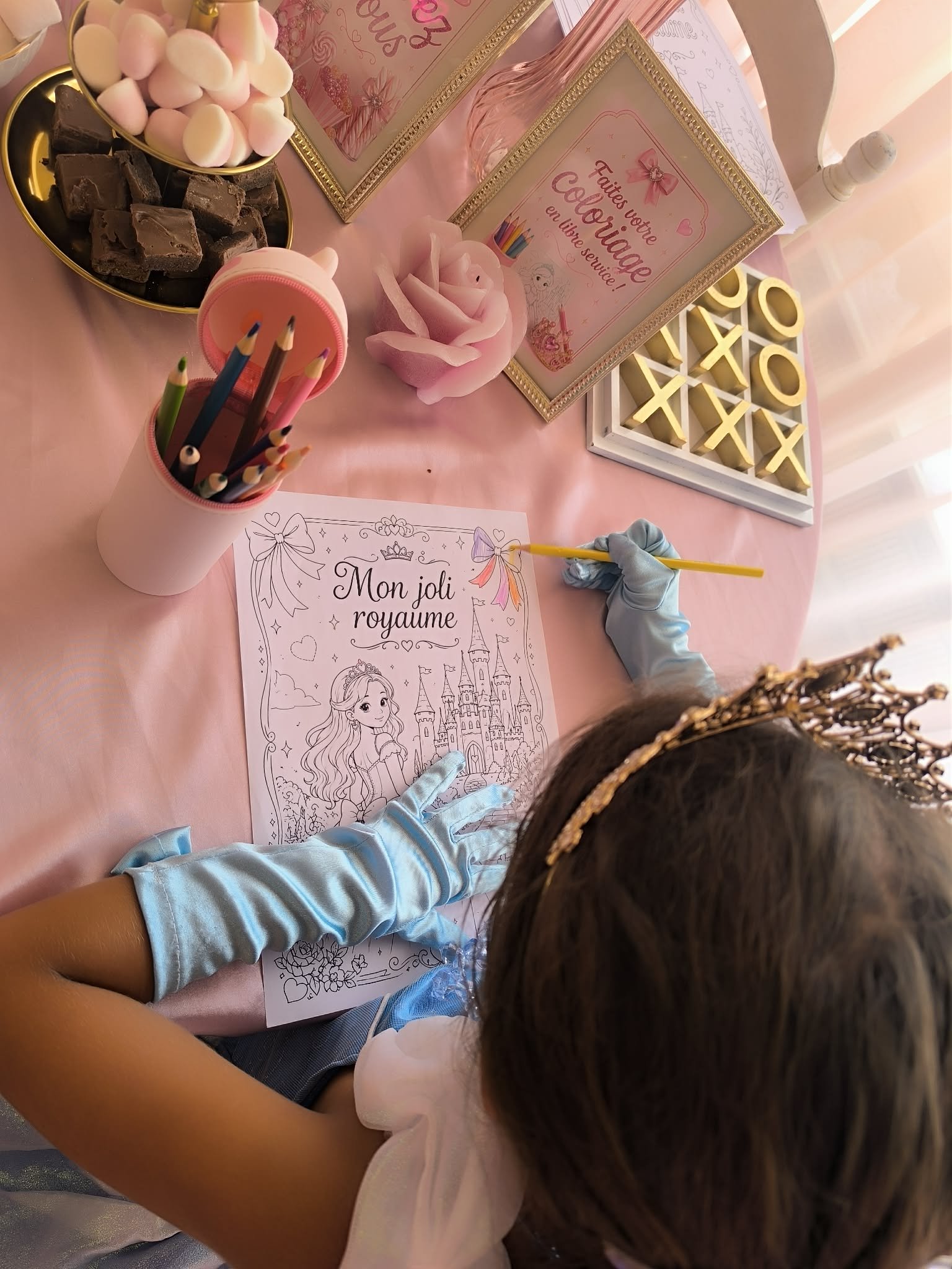 Une table rose avec des bonbons, des livres de coloriage pour filles, une rose en tissu, un jeu de morpion, et un cadre avec un message en français pour une fête fille ou un anniversaire.