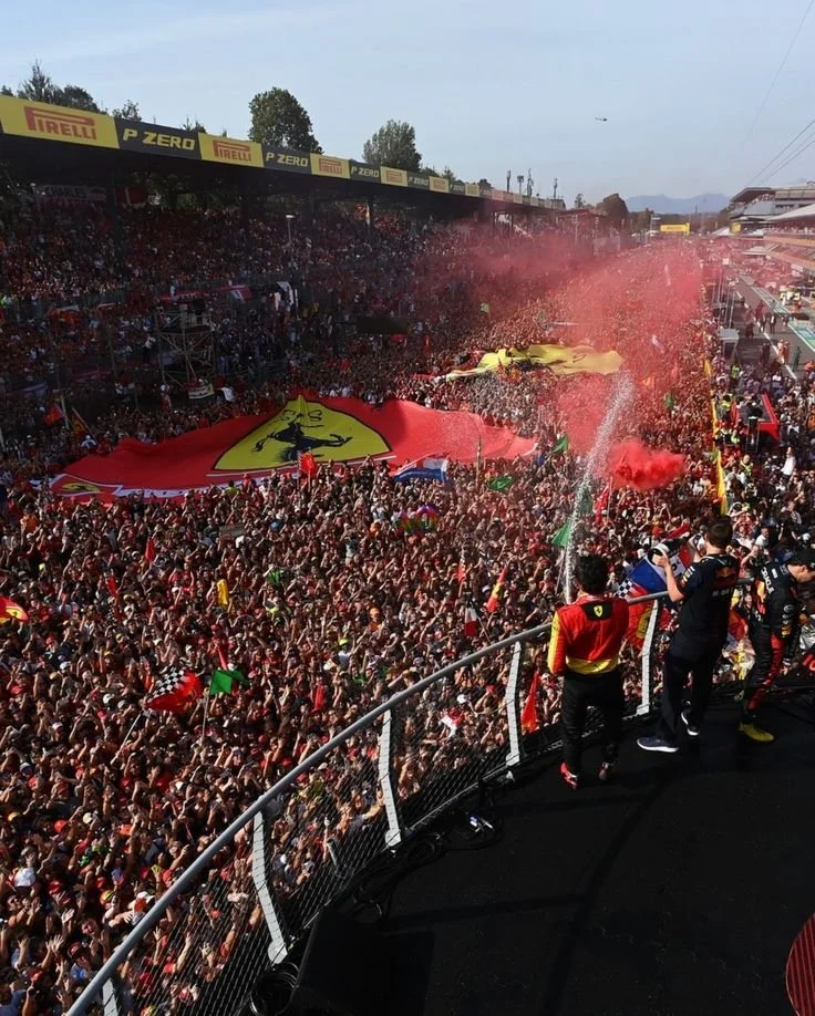 A large crowd of Ferrari fans at a racing event, waving flags, with a large yellow Ferrari banner in the background and red smoke in the air.