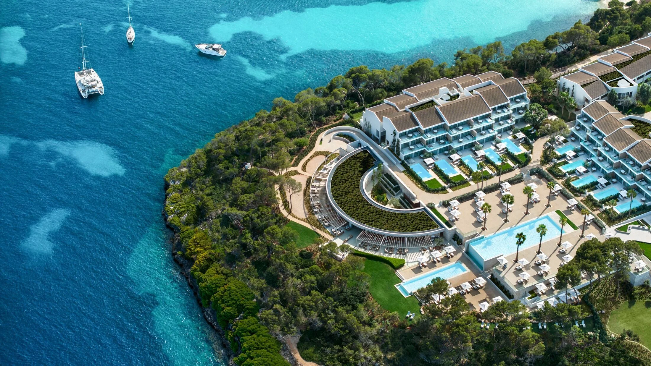 Aerial view of a beachfront resort with multiple buildings, swimming pools, palm trees, and boats anchored offshore in clear blue water.