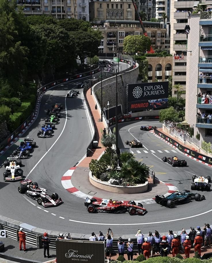 A group of race cars competing on a street circuit in Monaco, with spectators watching from the sides and balconies, surrounded by tall buildings and lush greenery.