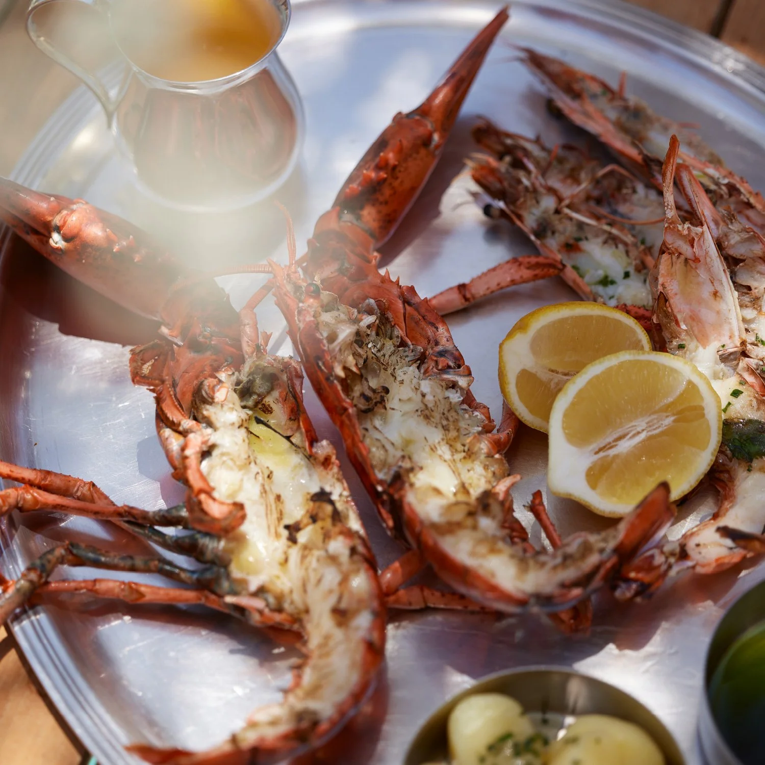 Lobster tails cut in half on a metal tray, garnished with lemon wedges, with a glass pitcher of melted butter nearby.