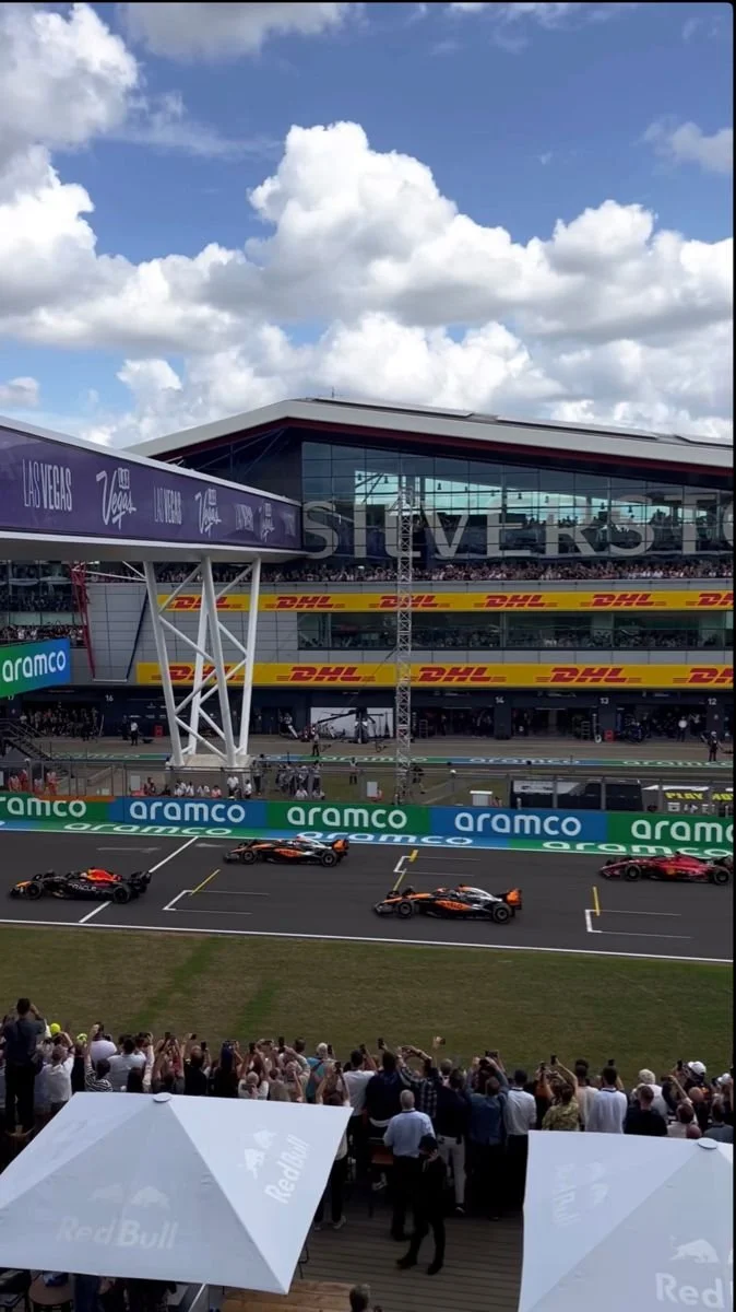 Formula race cars on the starting grid at a race track with spectators, sponsor banners, and a modern building in the background.