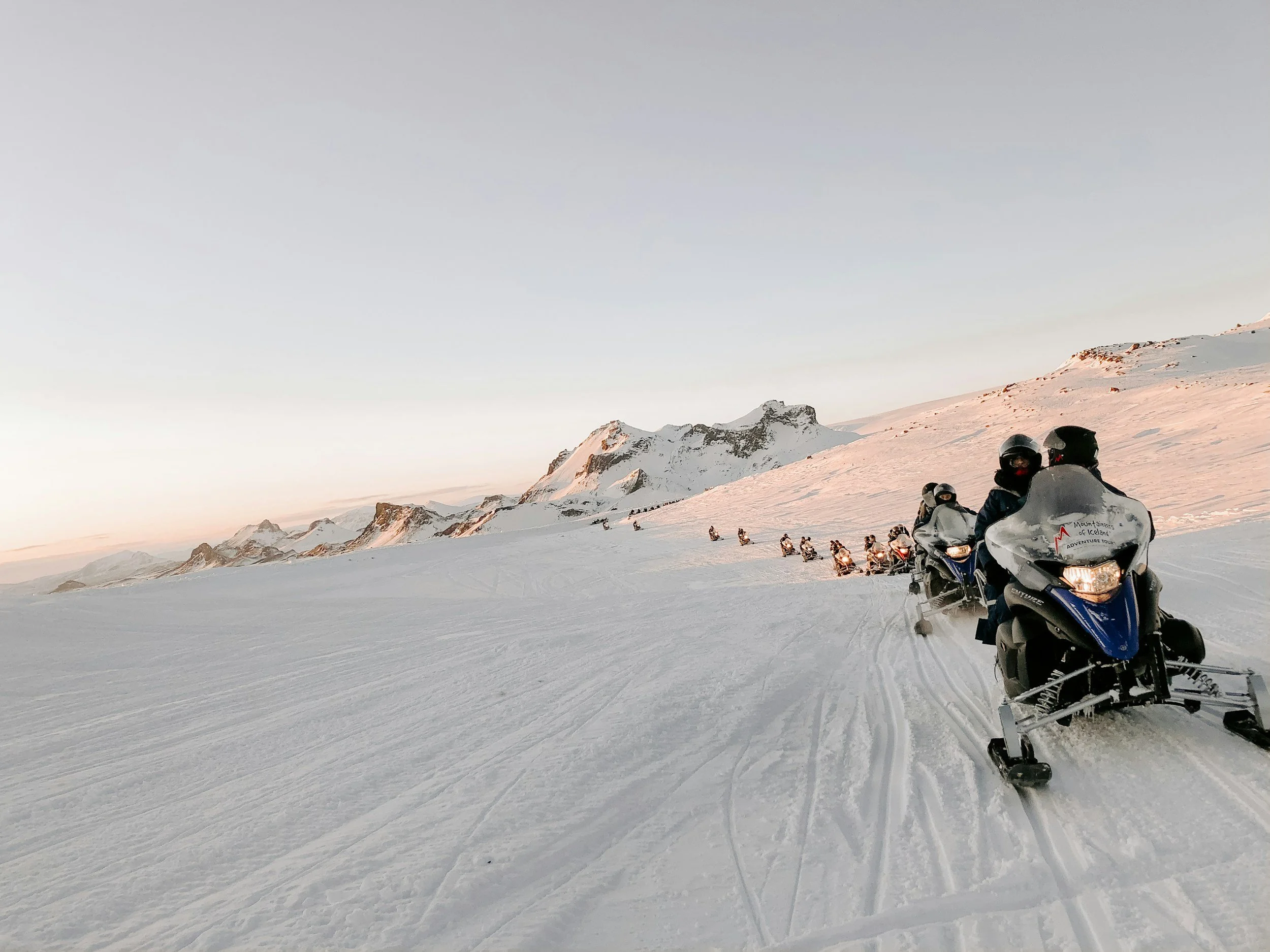 A line of people riding snowmobiles across snow-covered mountains at sunset.