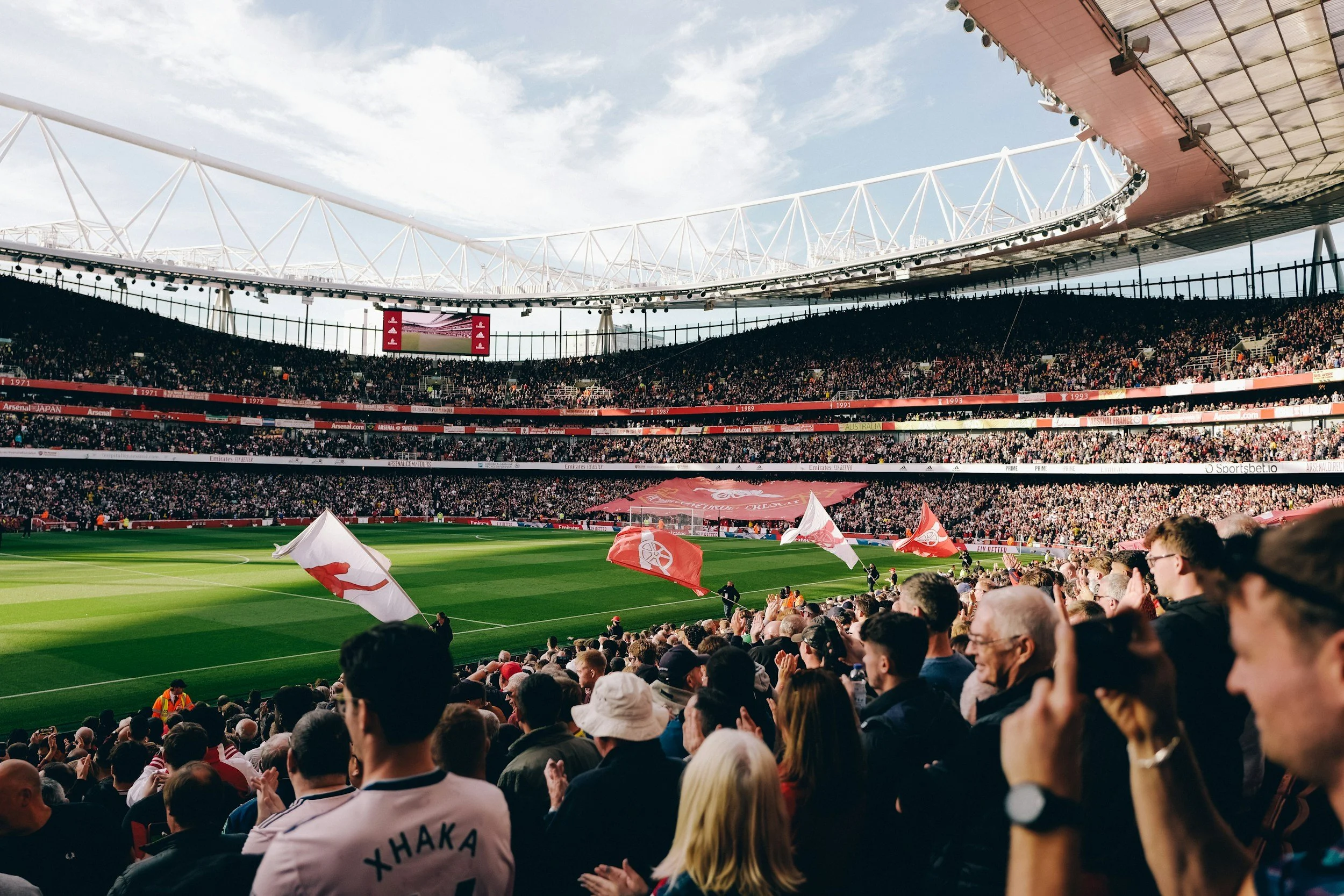 Crowd watching a soccer stadium filled with spectators, waving flags, with the field and stadium structure visible under a clear sky.