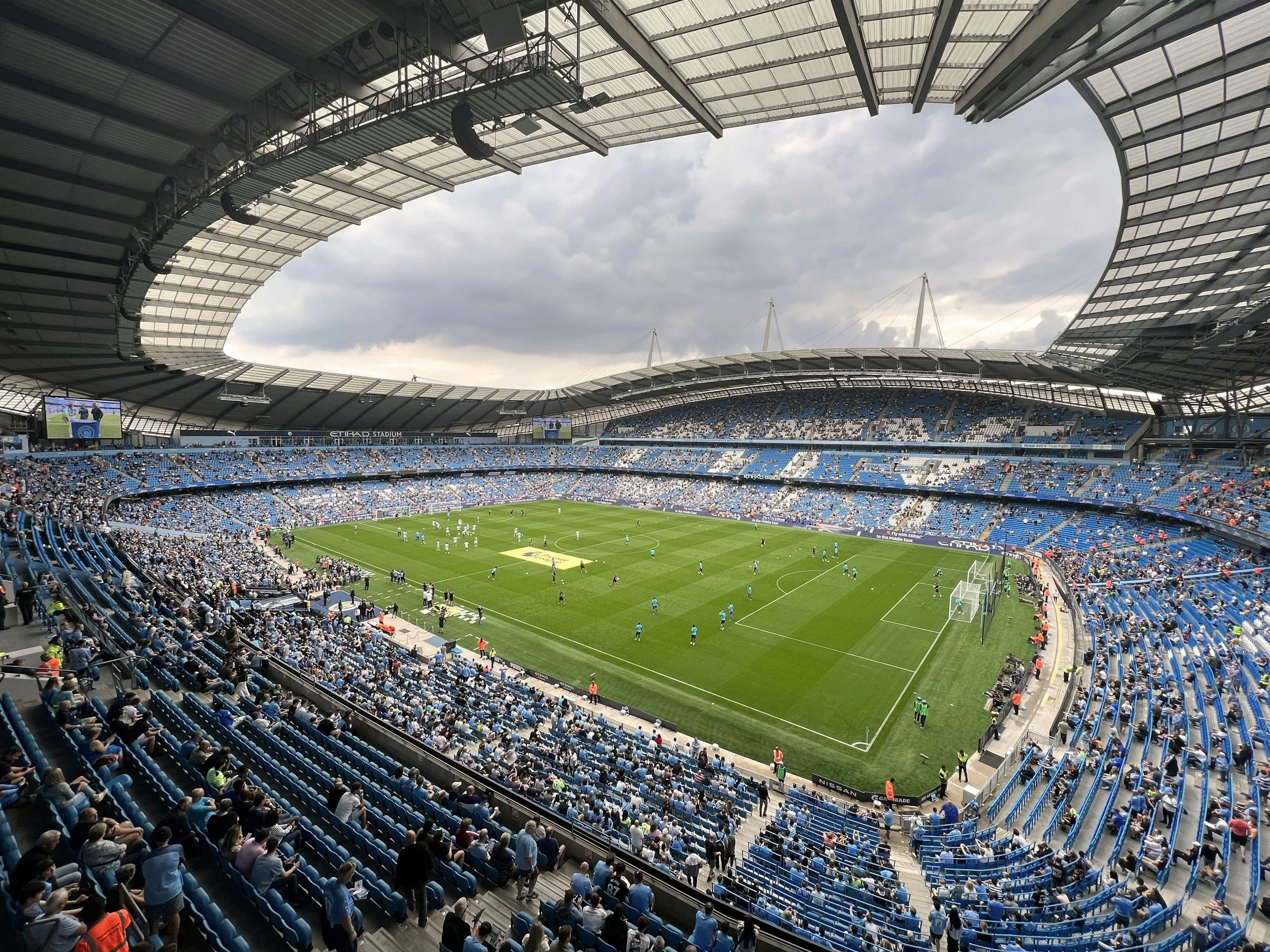 View of a soccer stadium with a green field, players warming up, and spectators seated in blue seats in a large open air stadium with a modern roof structure and cloudy sky.