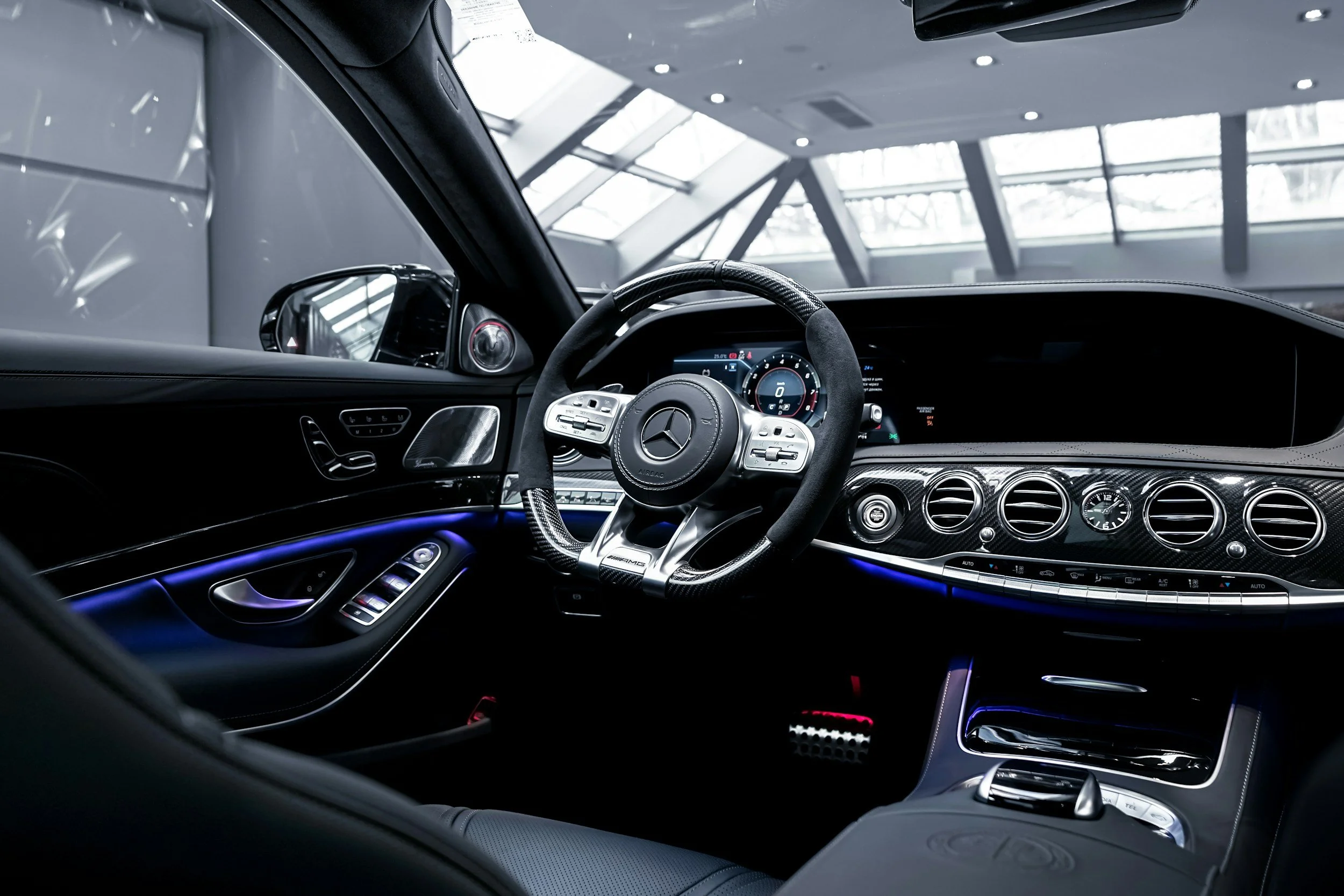 Interior view of a luxury Mercedes-Benz car dashboard with digital instrument cluster, carbon fiber accents, climate controls, and ambient lighting.
