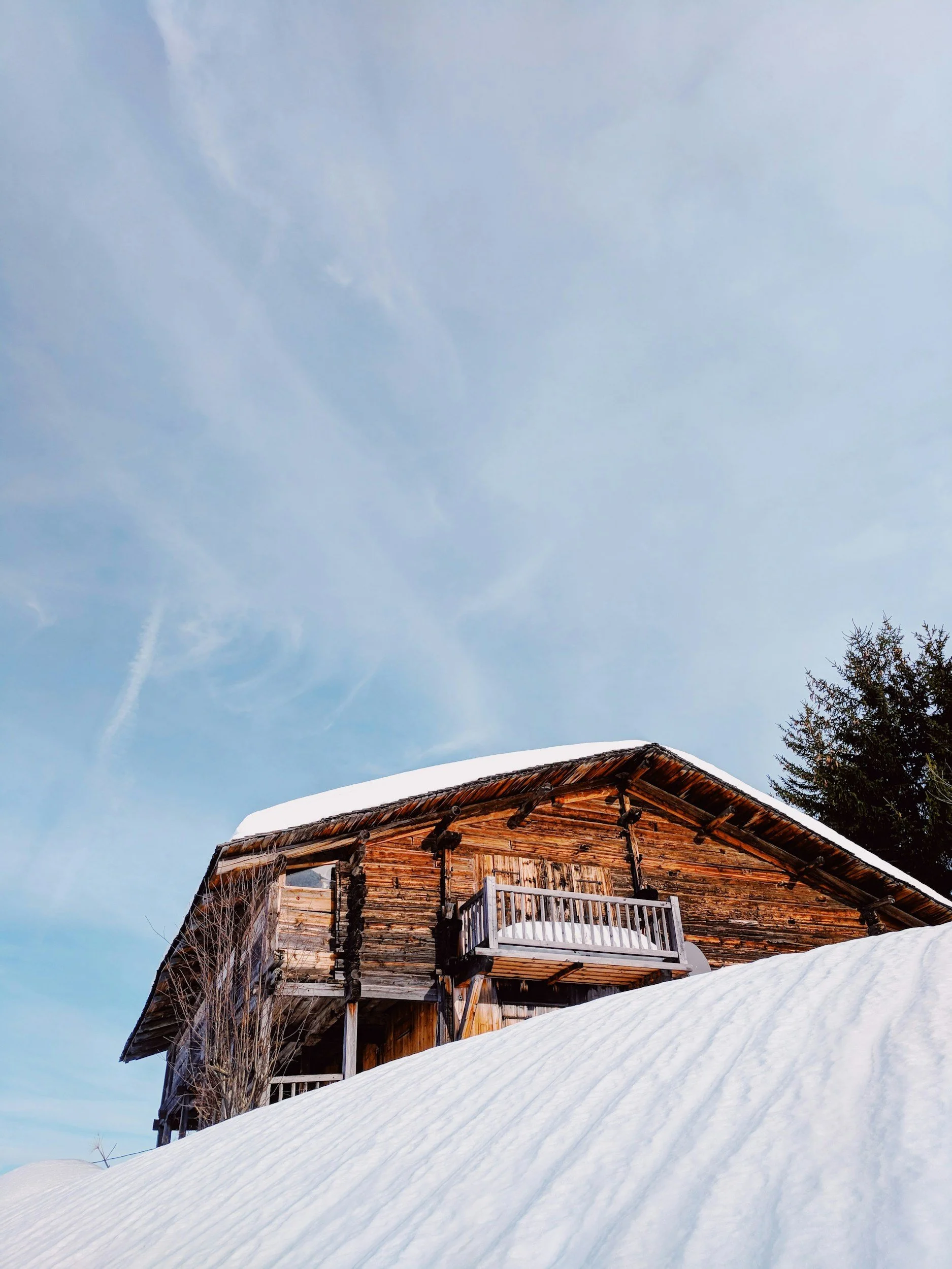 A rustic wooden cabin with a snow-covered roof, located in a winter landscape with snow on the ground and evergreen trees nearby, under a clear blue sky.
