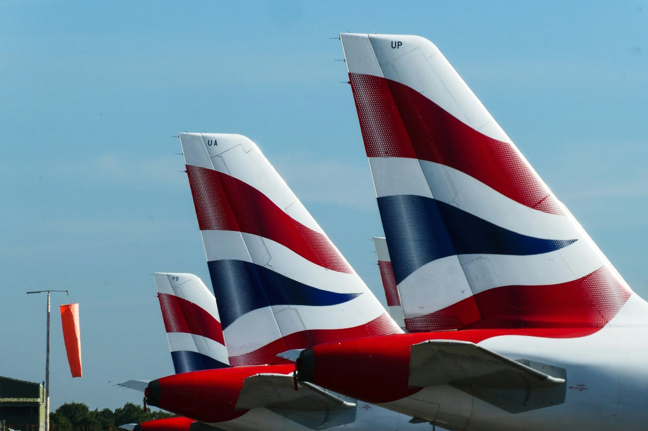 Close-up of the tails of three British Airways airplanes parked on the ground, showing their distinctive red, white, and blue tail designs against a blue sky.