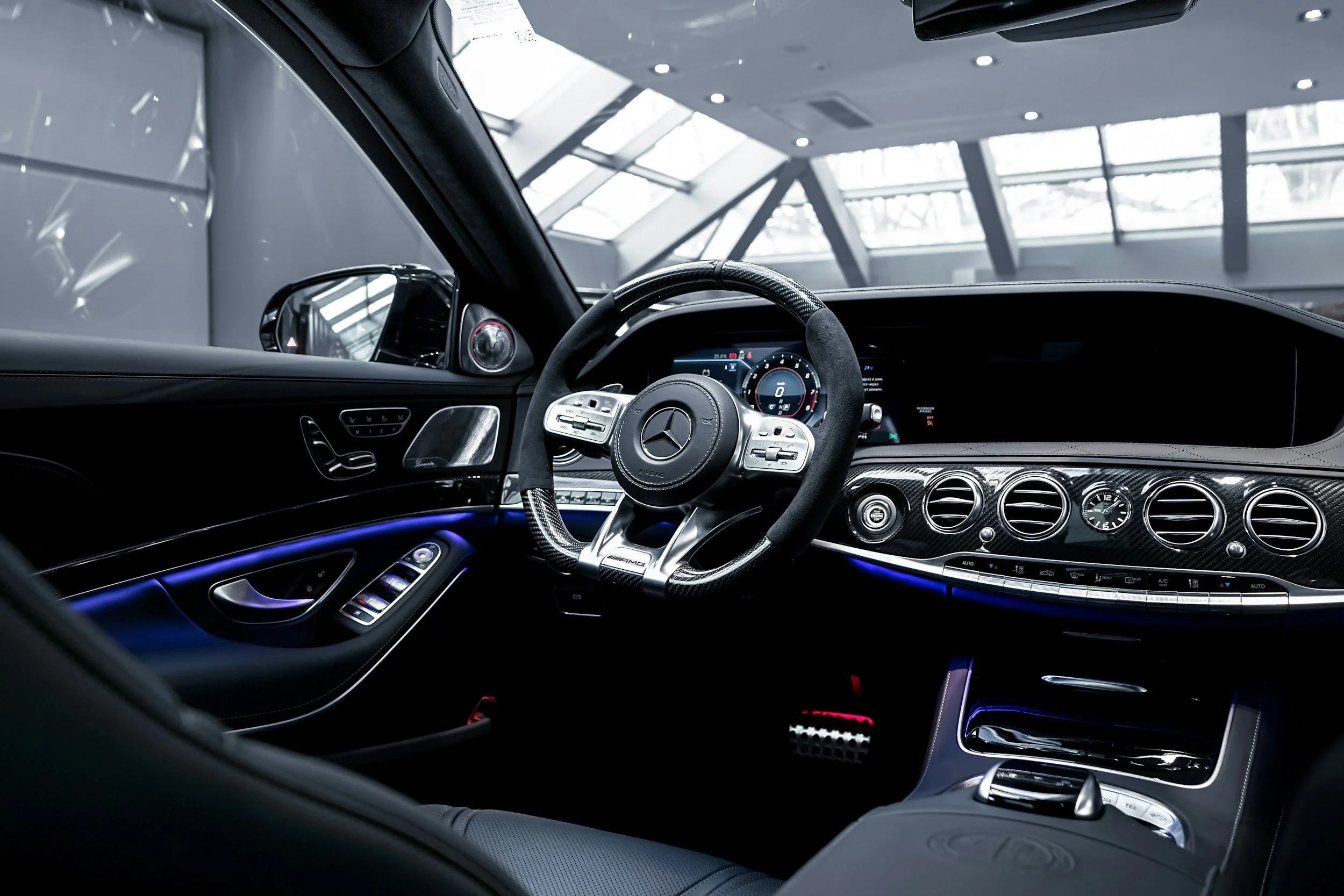The interior of a luxury Mercedes-Benz car, featuring a modern dashboard with digital displays, a steering wheel with Mercedes-Benz logo, carbon fiber accents, and blue ambient lighting.