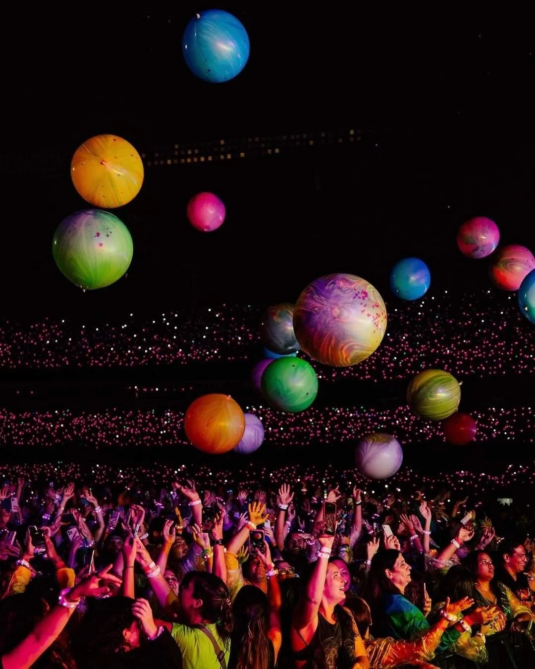 A crowd of people at a Coldplay concert, with many holding up their hands and smartphones. Above them, multicolored, glowing balloons or orbs float against a dark background, creating a vibrant and lively atmosphere.
