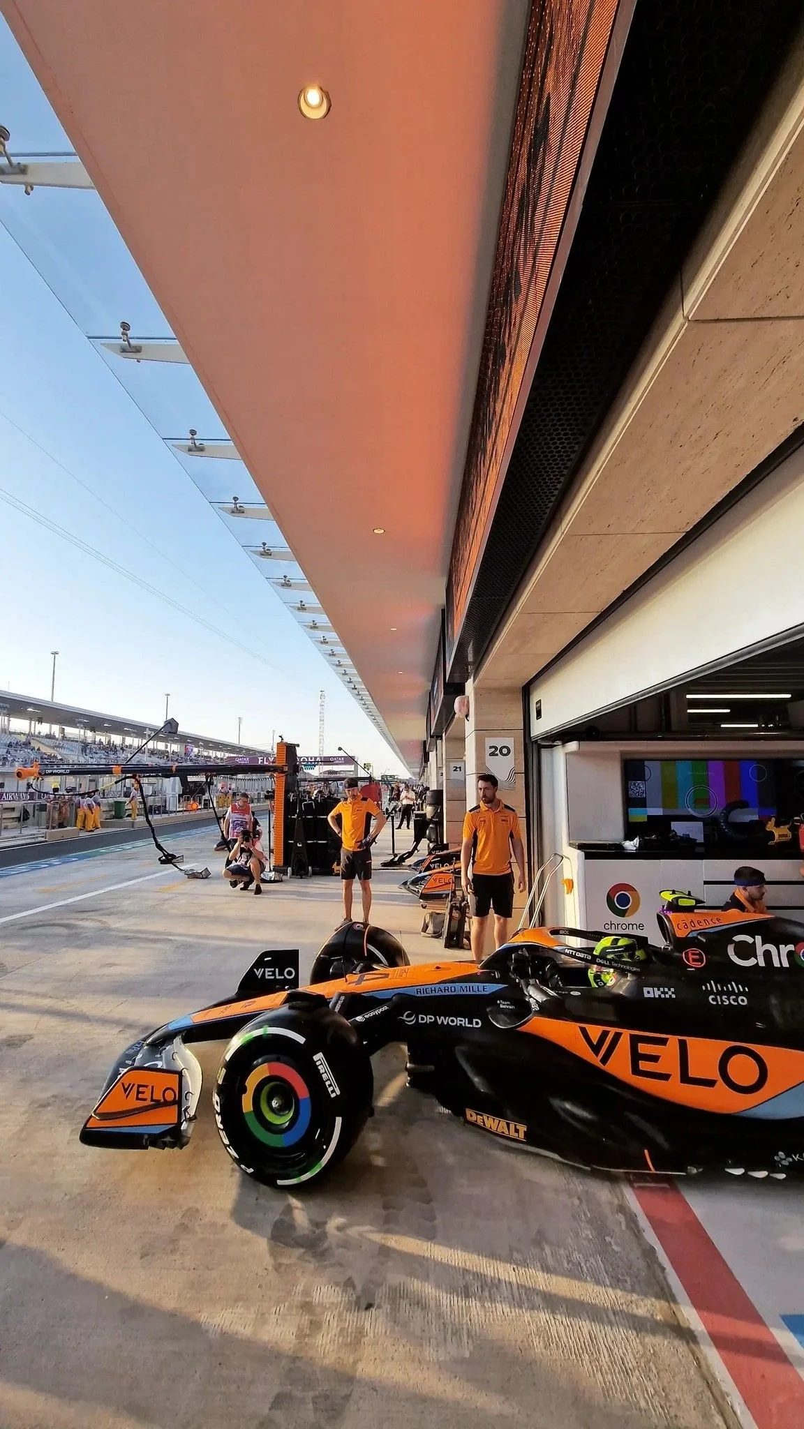 A Formula 1 race car in black and orange livery with sponsor logos, parked in a garage at a racing circuit during sunset with team members and equipment around.