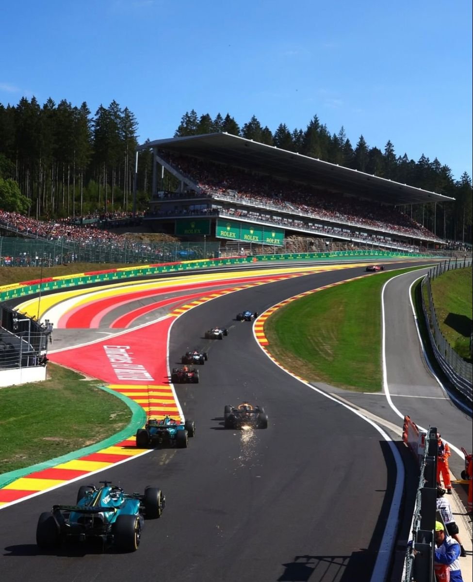 Cars racing on a race track with a grandstand and trees in the background.