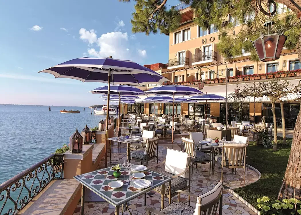 Outdoor waterfront restaurant with tables, chairs, and purple umbrellas, overlooking a large body of water with boats, next to a hotel with balconies.