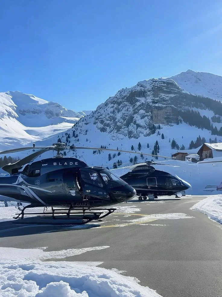 Two black helicopters parked on a snow-covered helipad in a mountainous area with snow-covered peaks and a clear blue sky in the background.