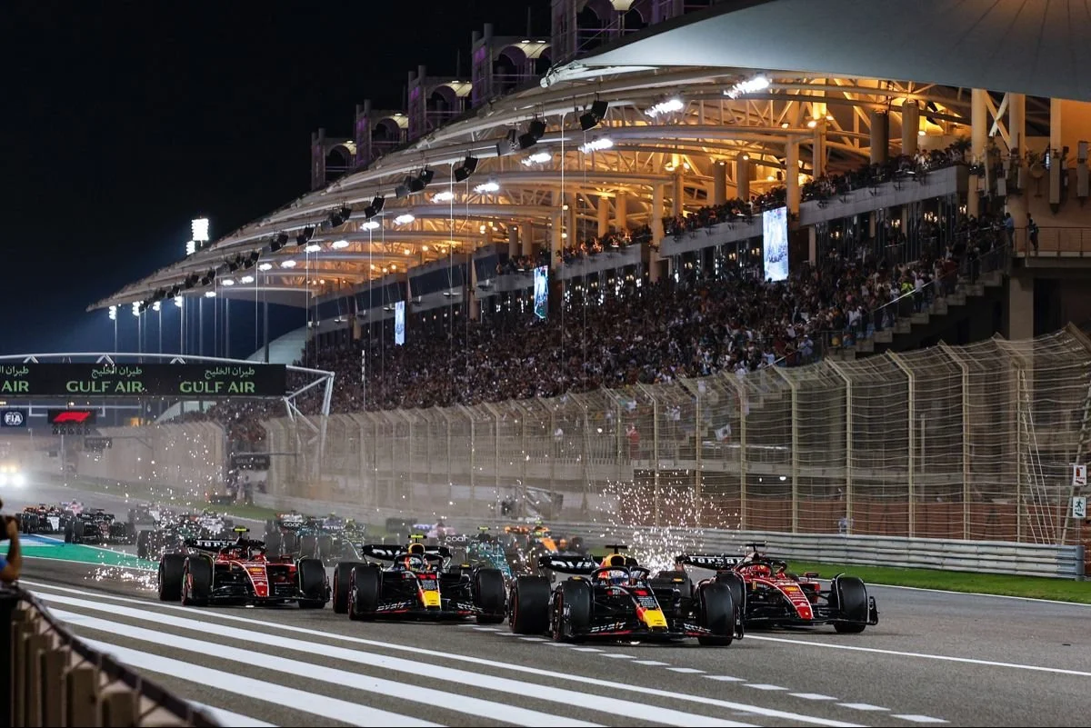 Nighttime Formula 1 race with multiple race cars driving on the track, sparks flying from the tires, spectators in the grandstands, and bright lights illuminating the scene.