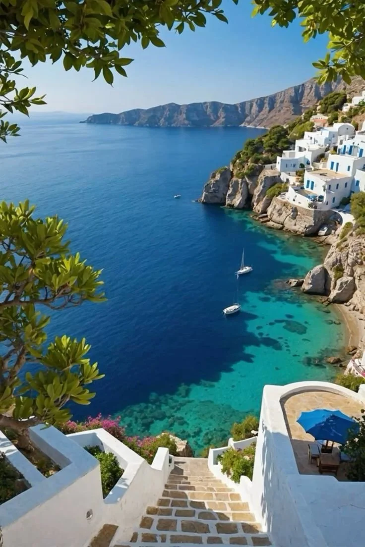 Aerial view of a coastal village with white buildings on rocky cliffs, clear blue water with boats, and a staircase leading down to the beach.