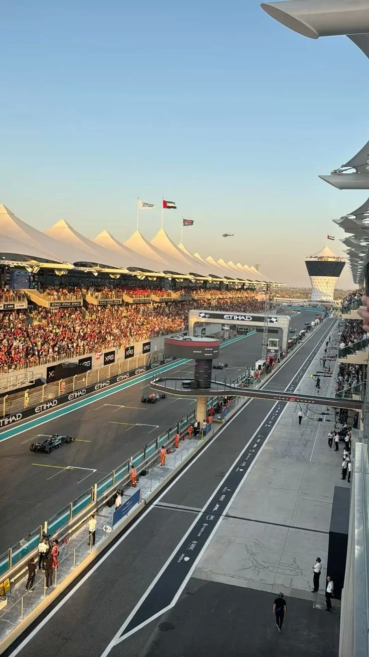 View of a Formula 1 race track at sunset with racing cars, spectators in the grandstands, and team personnel near the pit lane.