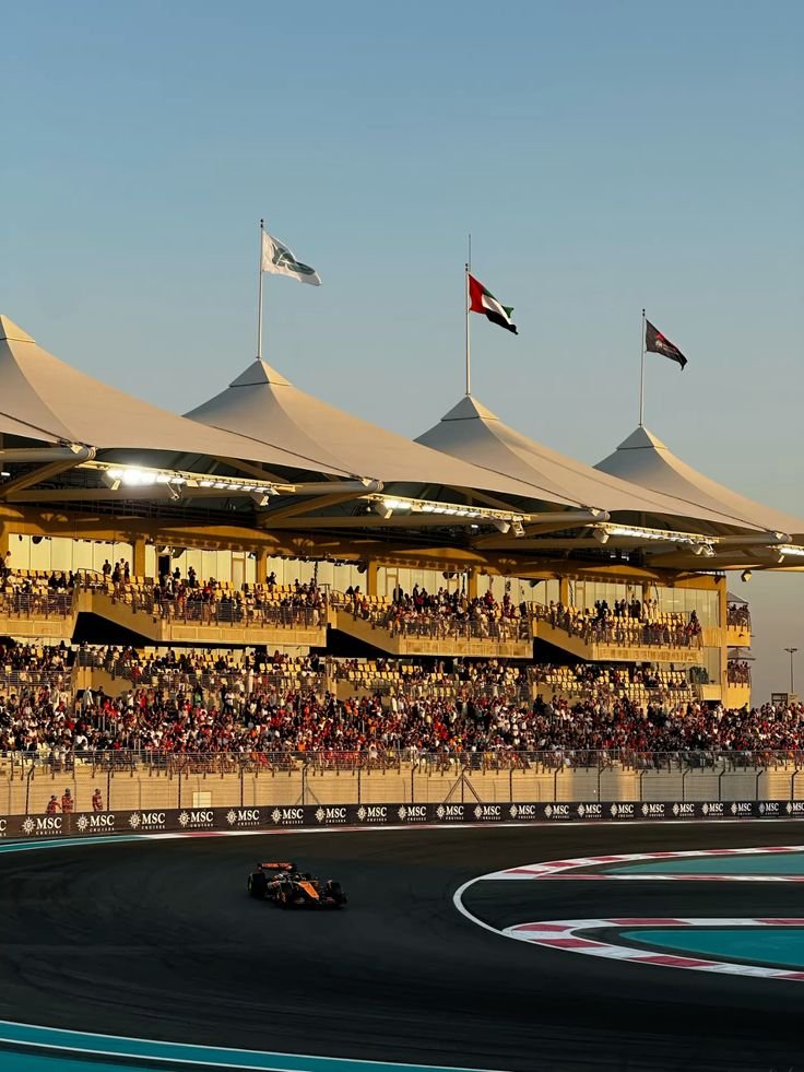 A race car driving on a racetrack in front of grandstand filled with spectators, with a modern building featuring flags on top in the background.