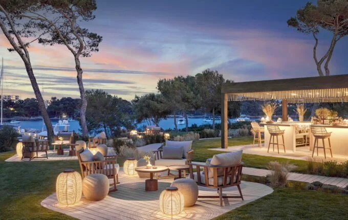 Outdoor lounge area with wooden chairs, cushions, and lanterns, overlooking a marina at sunset.