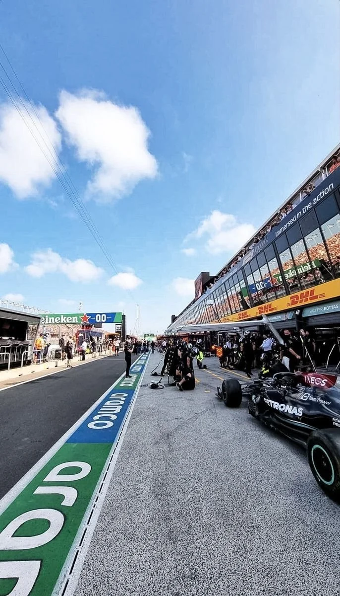 Formula 1 race car in the pit lane with crew members working on it, race track with sponsors' signage, and spectators in the background under a partly cloudy sky.