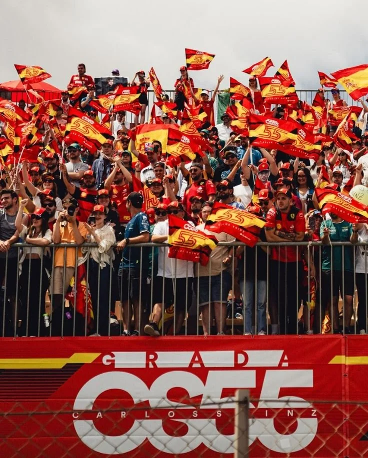 Crowd of people celebrating with red and yellow flags at a race event, with a banner that says 'GRADA OSCAR CLOSSINZ' in front.