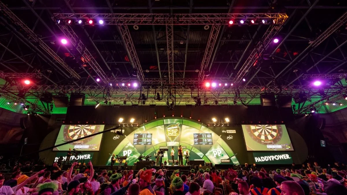 An indoor stage at a professional darts tournament with bright, colorful lighting and two large screens displaying scores. The audience is seated in front, with some wearing hats and colorful attire.