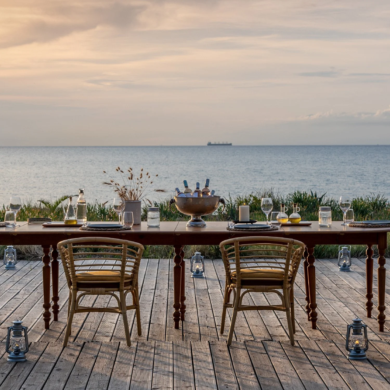 A seaside outdoor dining table set for a meal at sunset with chairs, glasses, bottles, flowers, and lanterns, overlooking the ocean and a ship in the distance.