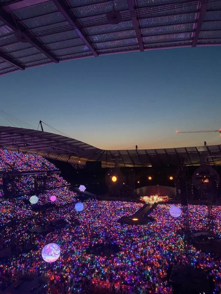 Large outdoor concert stadium filled with thousands of people holding colorful glow sticks at sunset, with a stage in the distance and a partially covered roof overhead.