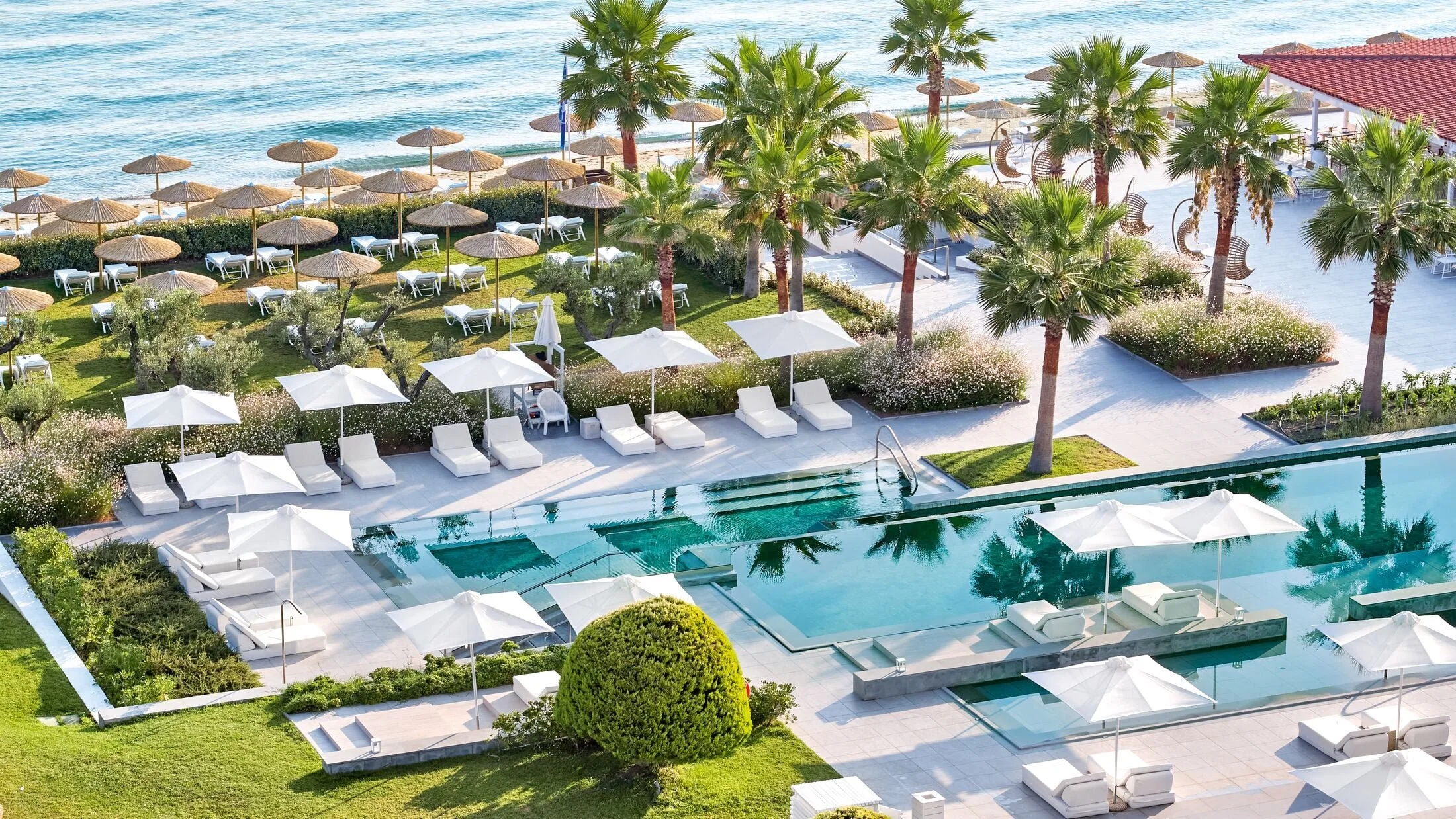 Resort pool area with white lounge chairs, umbrellas, and palm trees near the ocean.
