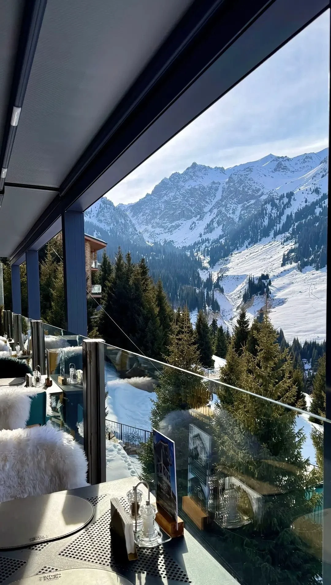 Indoor dining area with a view of snow-covered mountains, trees, and a partly cloudy sky outside a restaurant with glass walls.