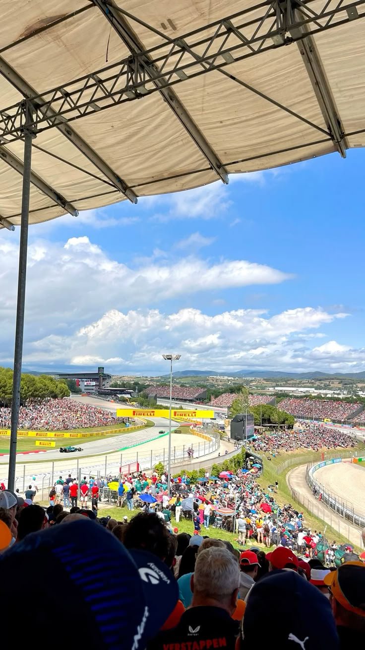 Spectators in a race track stadium watching a Formula 1 race with a sunny sky, clouds, and mountains in the background.