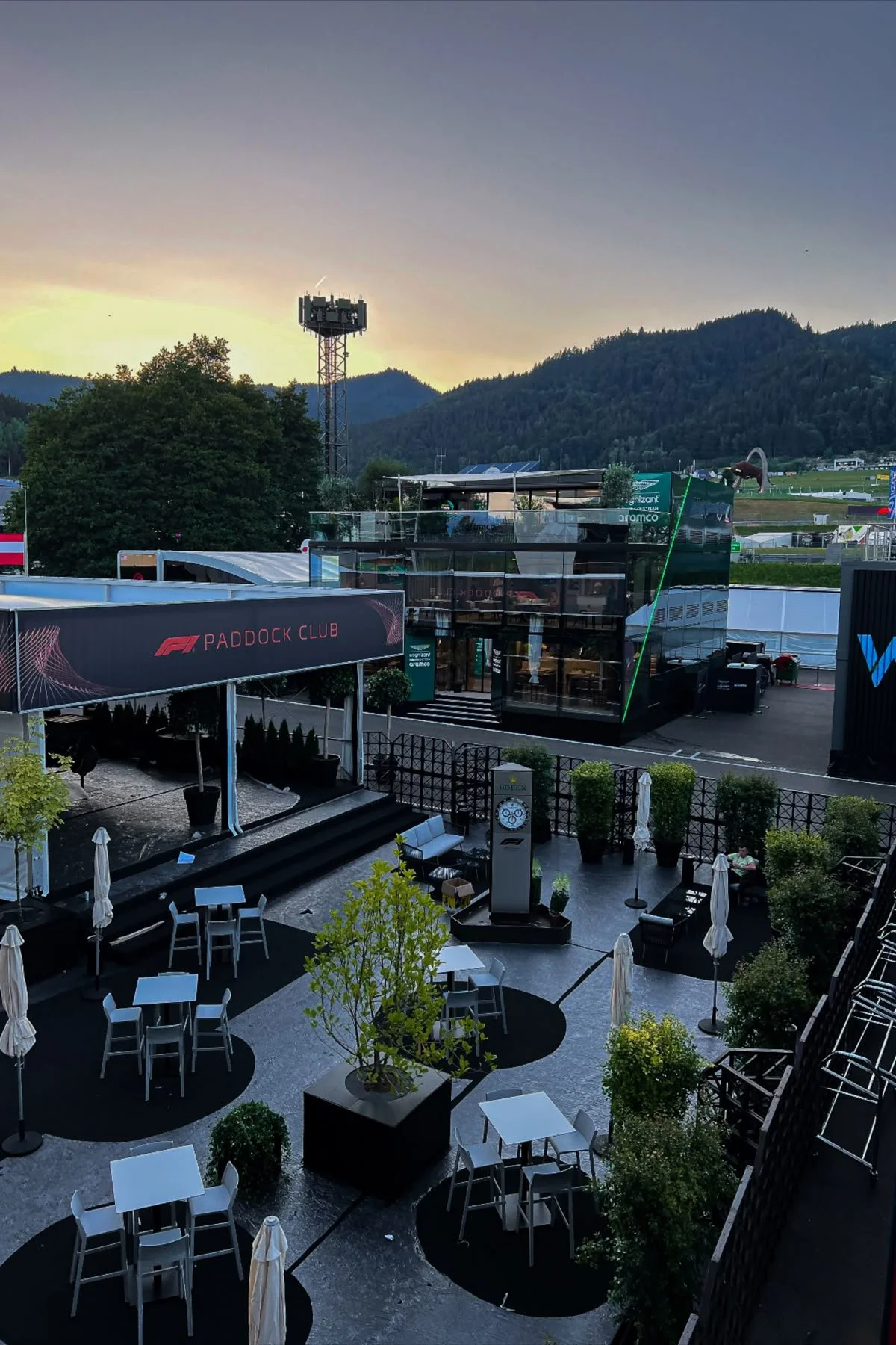 Empty outdoor patio with white tables and chairs, umbrellas, and potted plants at a racing event venue during sunset.