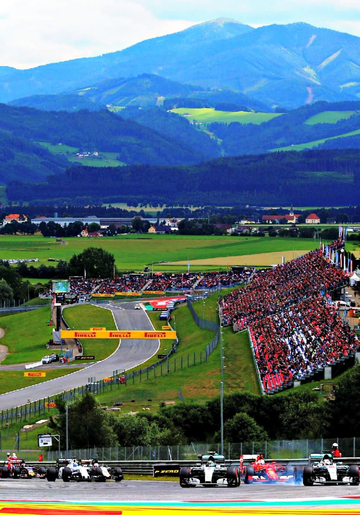 Formula 1 race at a track with a spectator stand full of people, surrounded by green fields and mountains in the background.