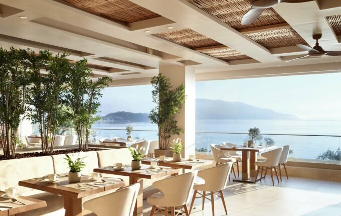 Indoor restaurant with large windows overlooking water and mountains, decorated with plants, white chairs, and wooden tables.