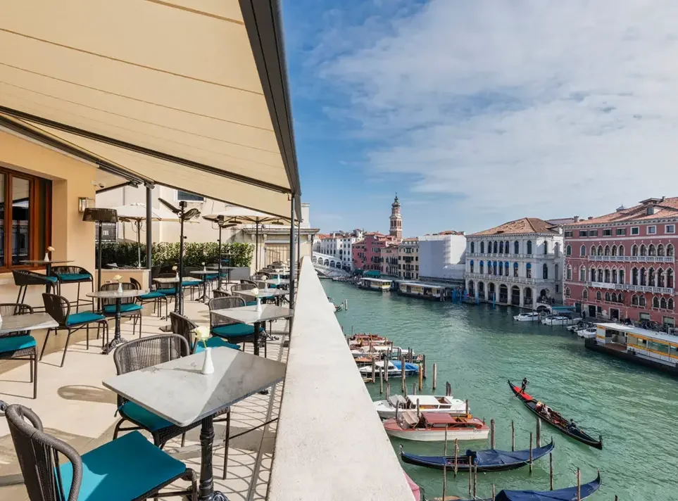 Outdoor restaurant patio overlooking a canal with boats in Venice, Italy, on a sunny day.