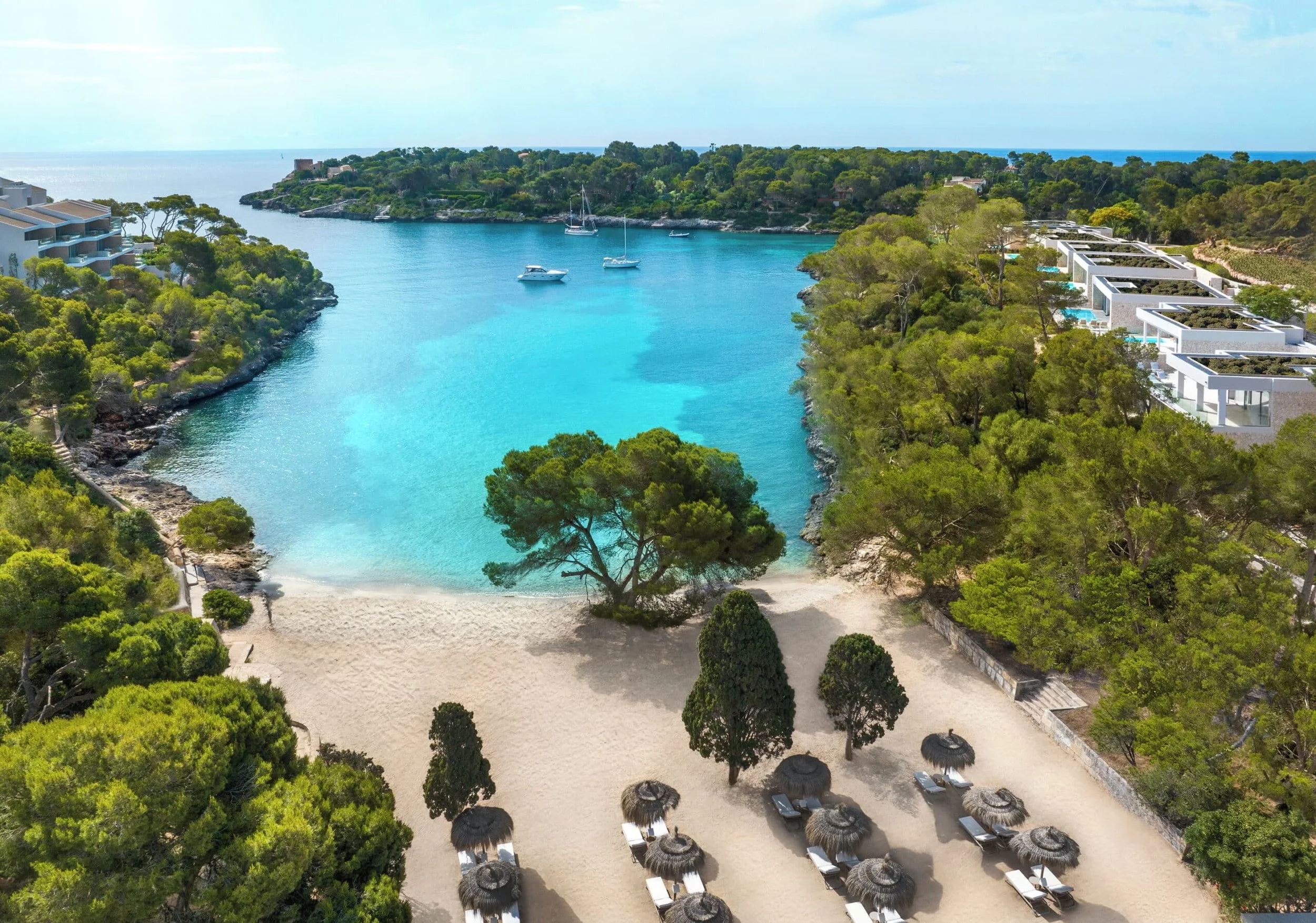 A scenic view of a secluded beach with white sand, surrounded by lush green trees, with a boat-filled blue water bay in the background. There are cabanas with thatch roofs and sun loungers on the beach.