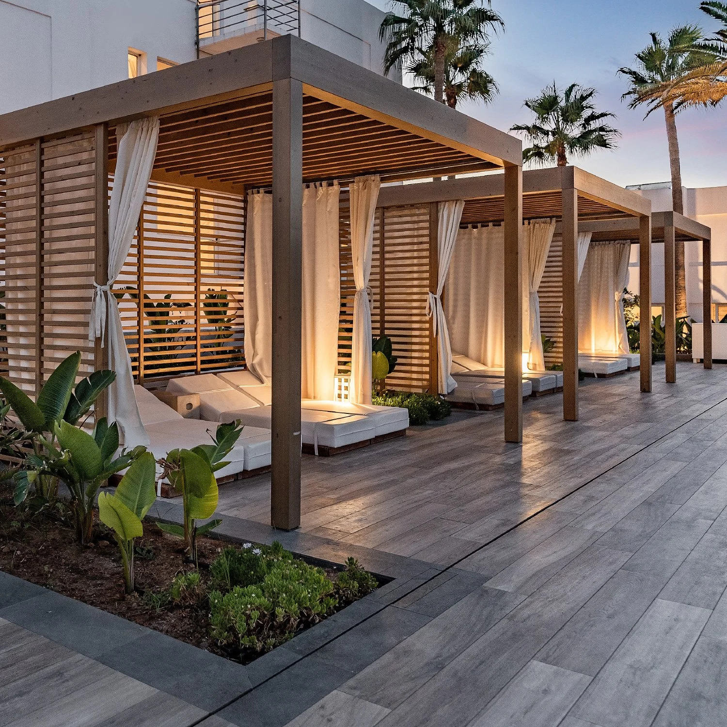 Outdoor lounge area with wooden cabanas, white curtains, and cushioned loungers illuminated by soft lighting, with palm trees in the background during sunset.