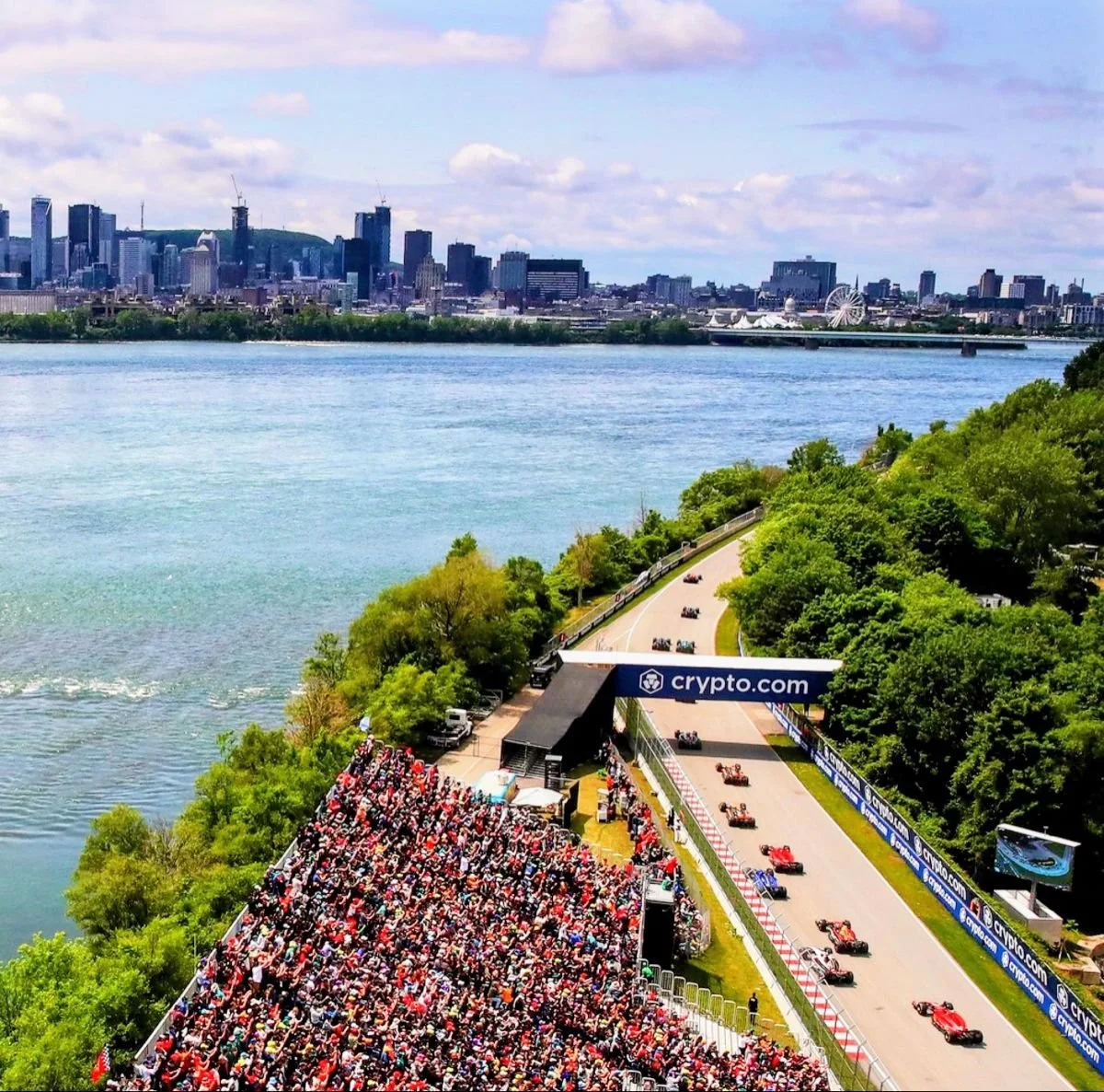 A race track along a river with a large crowd of spectators in the grandstands and racing cars on the track, with a city skyline and a Ferris wheel in the background.