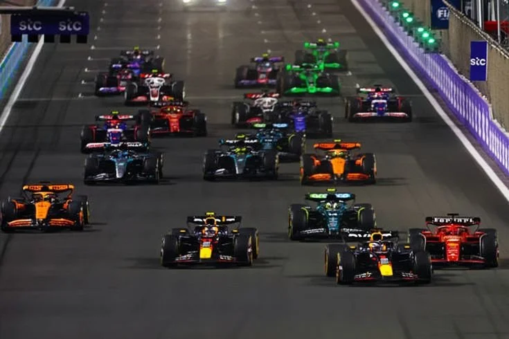 Multiple Formula 1 race cars on a racetrack during a race, lined up and approaching the finish line, with some cars in the lead and others behind, under bright racing lights.