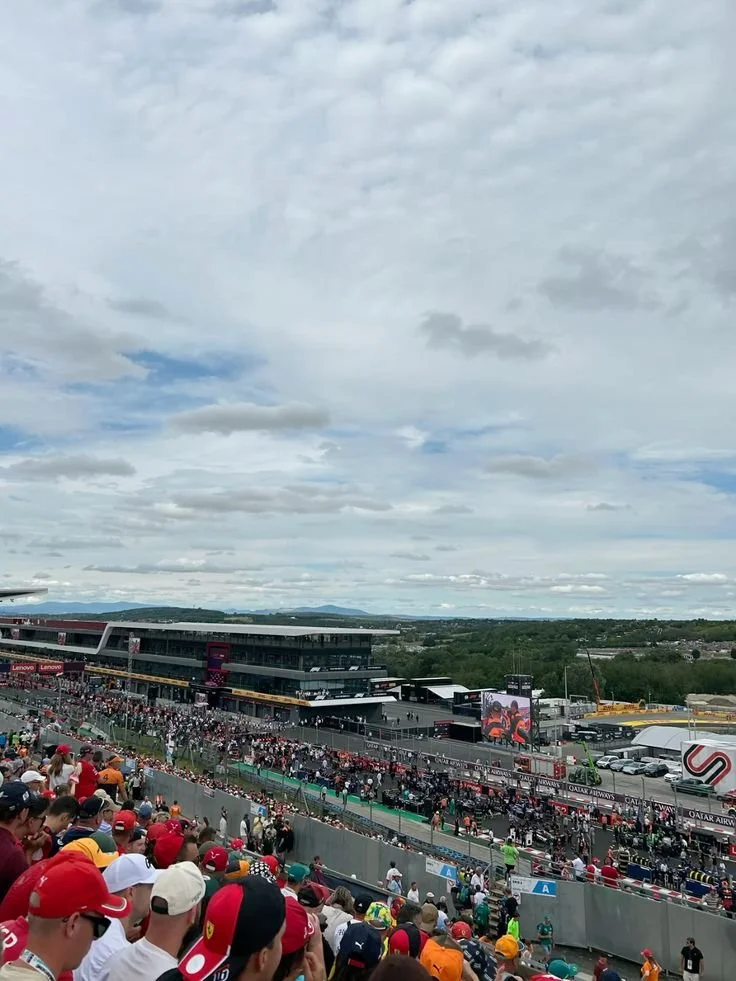Crowd at a racing event with race cars on track and a large screen displaying race footage.