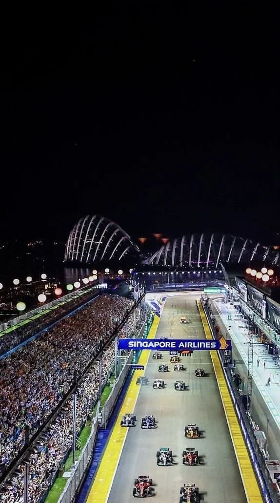 Nighttime view of a Formula 1 race track with racing cars on the starting grid, a large crowd of spectators, and the Marina Bay Sands hotel in Singapore in the background.