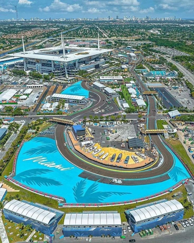 Aerial view of the Miami Grandstand at the Miami Grand Prix with a street circuit featuring colorful track walls, palm tree designs, and a large stadium in the background.