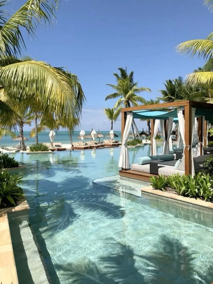 Luxury resort pool area with palm trees, lounge chairs with white umbrellas on the beach, and a cabana with cushions, overlooking the ocean under a clear blue sky.
