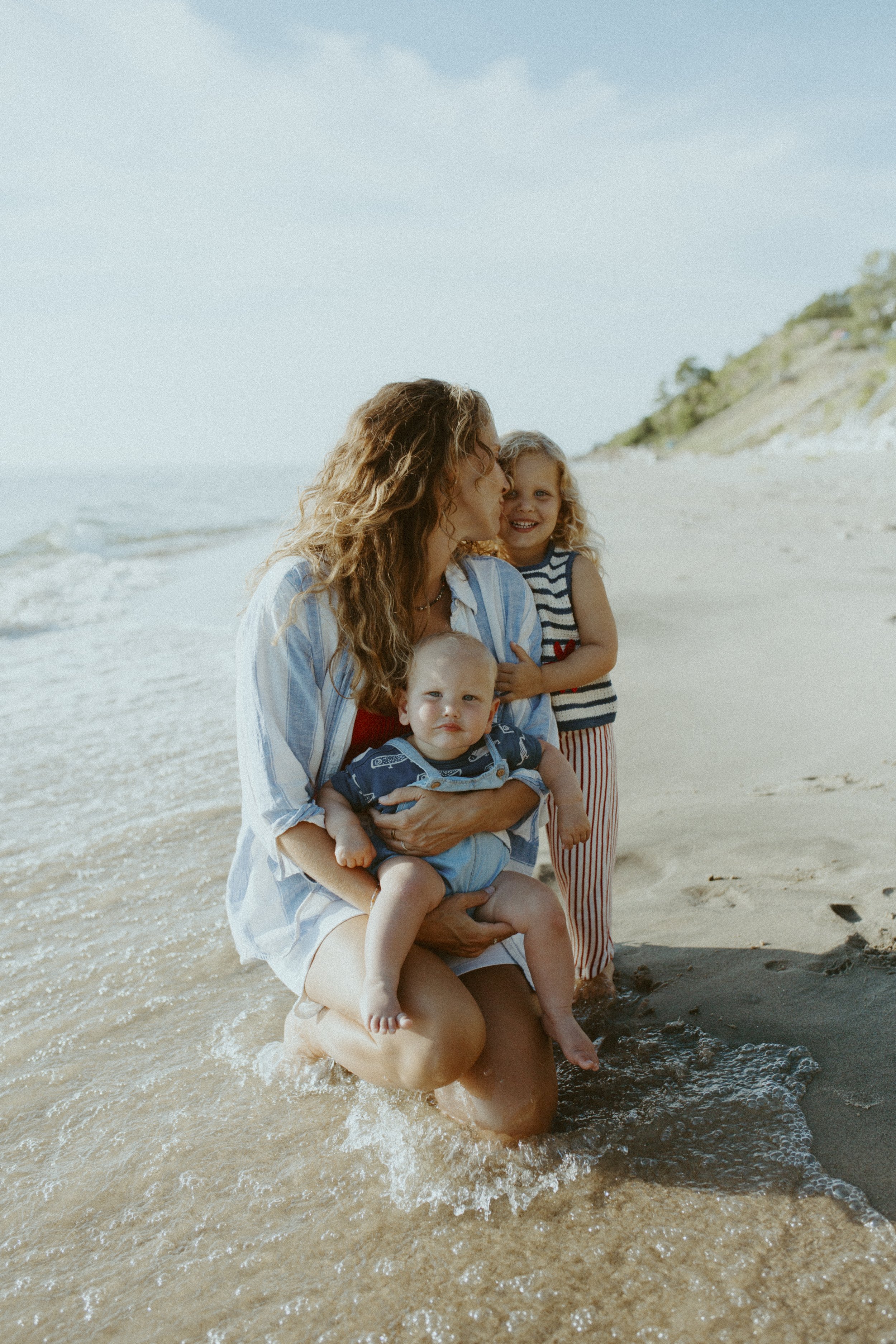 A woman with long curly hair holding two young children, one boy and one girl, on a beach with waves and a hillside in the background.