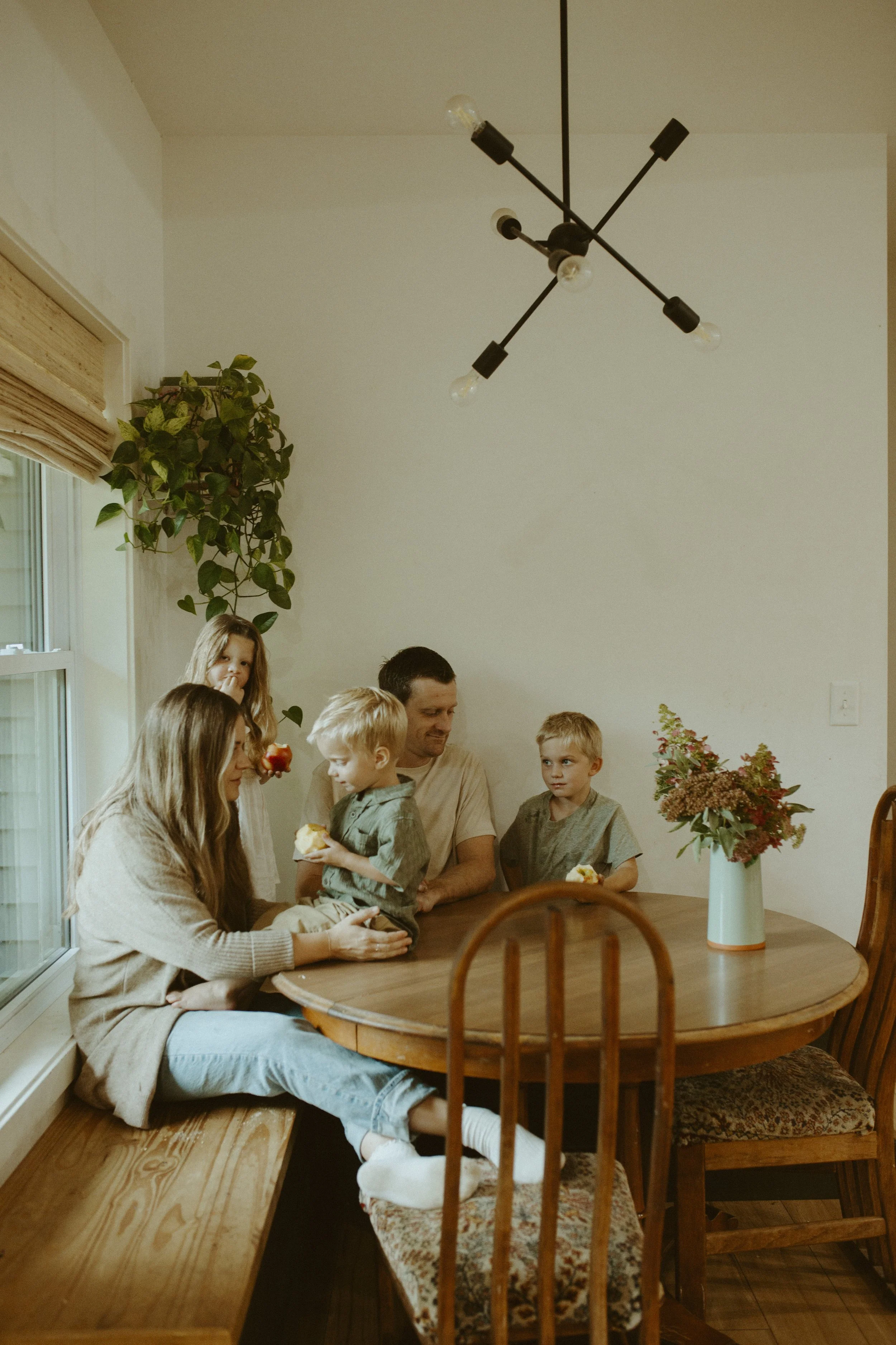 Family gathered around a wooden dining table with a vase of flowers, some children and adults eating and talking inside a cozy dining room with a hanging light fixture and windows with blinds.