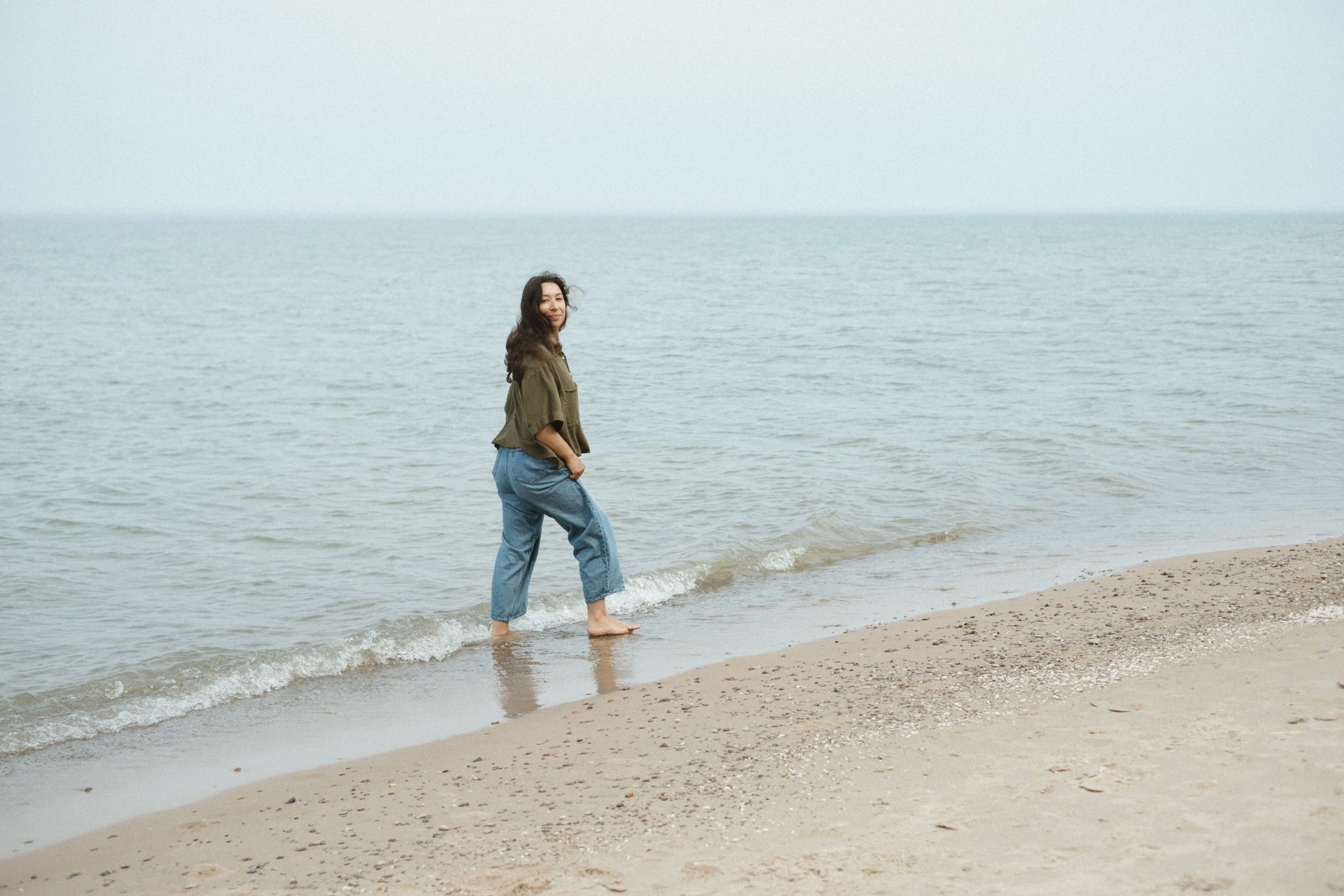 A woman with long dark hair wearing a green jacket and blue jeans standing in shallow water at the beach, facing the camera with the ocean and sky in the background.