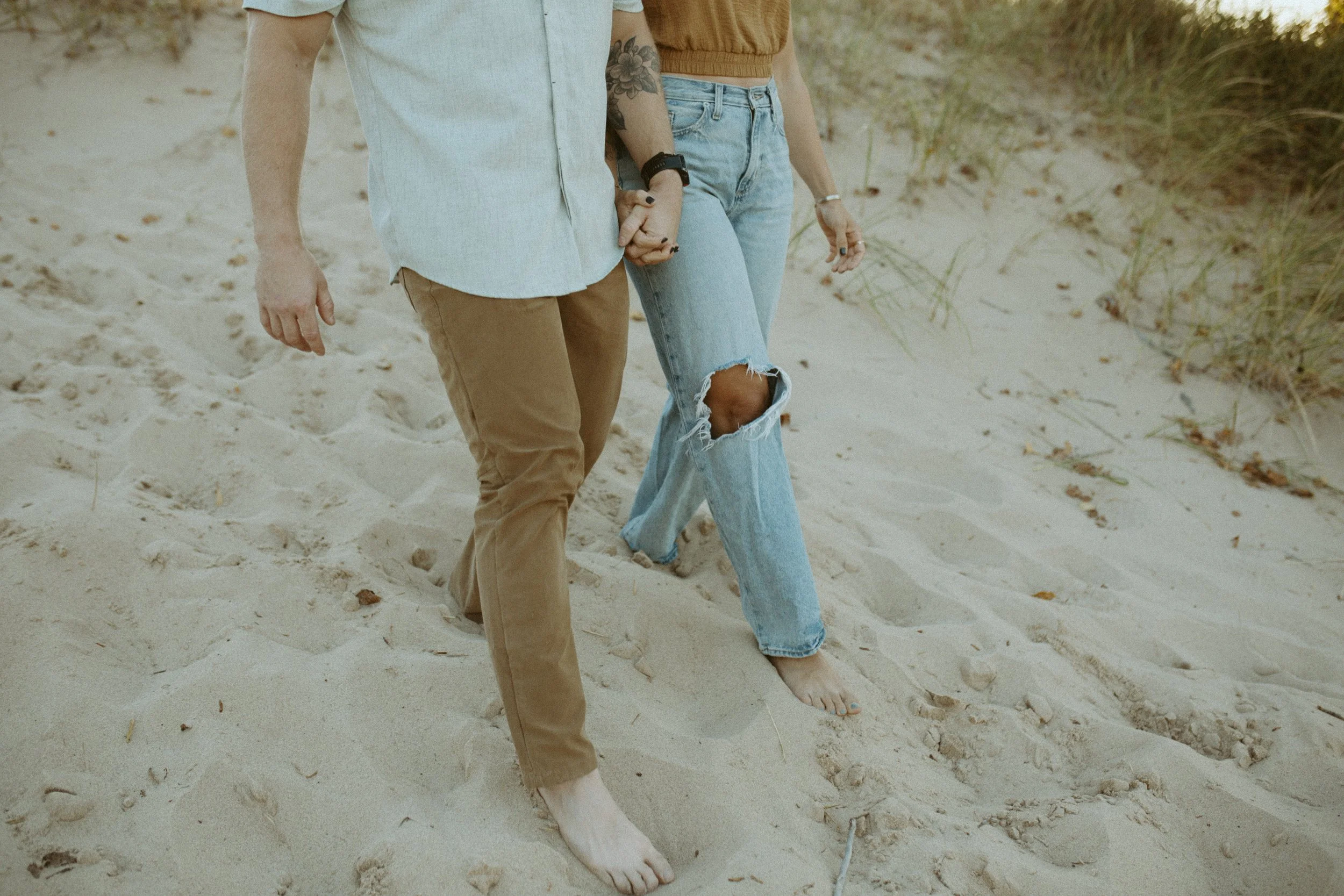 A couple holding hands while walking barefoot on the sand at the beach, with grass and dunes in the background.
