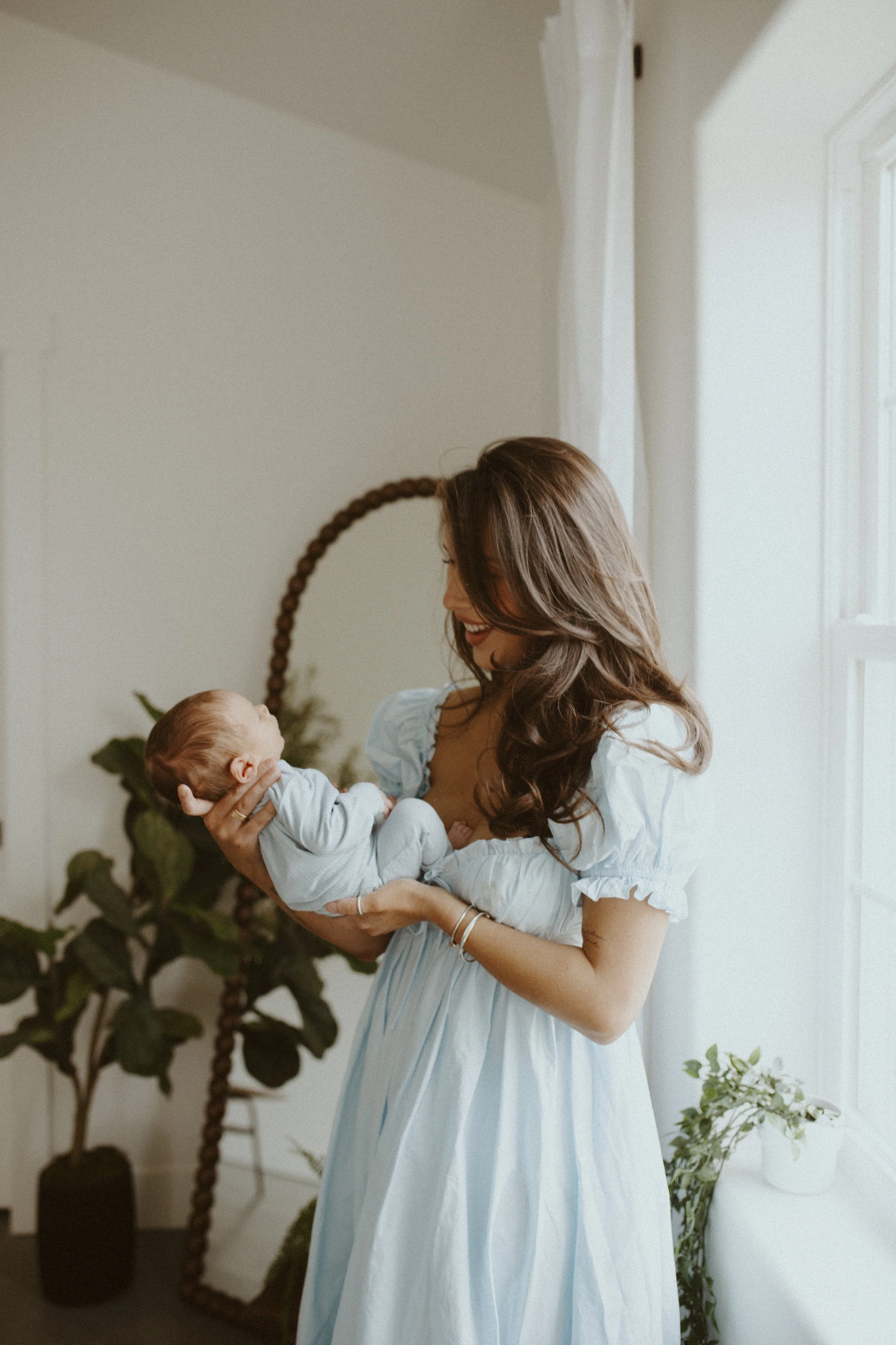 A woman with long, wavy brown hair wearing a light blue dress holding a newborn baby in a bright, naturally-lit room with plants and white walls.